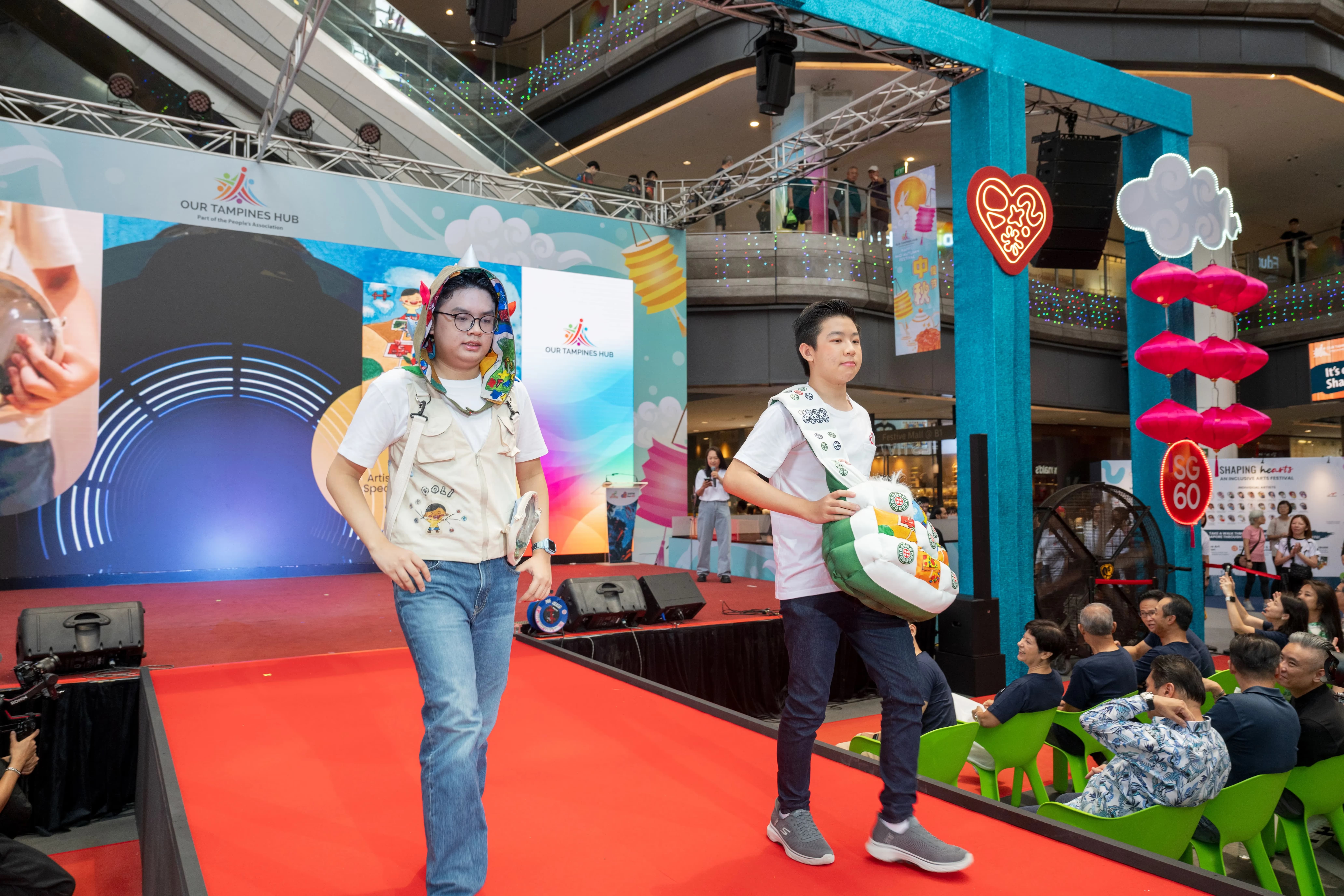 Persons with disabilities walking on the fashion runway, carrying bags printed with artwork by Shaping Hearts artists.